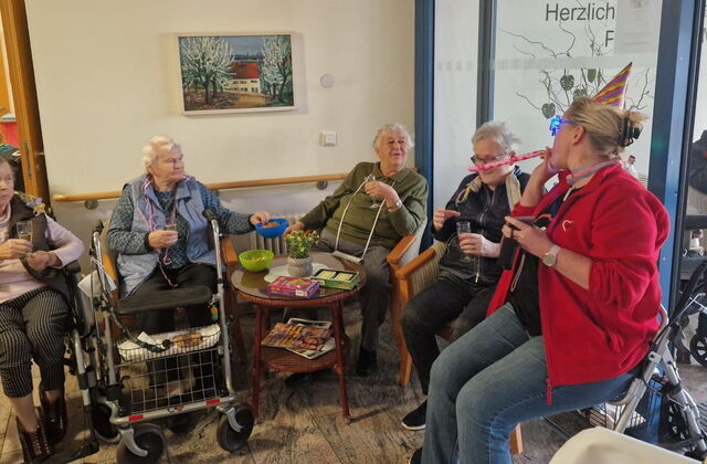 4 Seniorinnen und einer Betreuerin in einer Sitzecke. Die Betreuerin trägt ein spitzes Faschindhütchen bläst in bunte Fasching-Pfeife. Fröhliches Beisammensein.