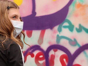 Frau mit weißen Handschuhen und OP-Maske, Sie greift nach eine Spraydose. Vor ihr eine besprühte Graffiti Wand.
