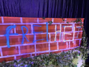 Bühnenbild. Eine Ziegesteinmauer von Efeu umrangt und mit Graffiti ist das Wort: FREIHEIT