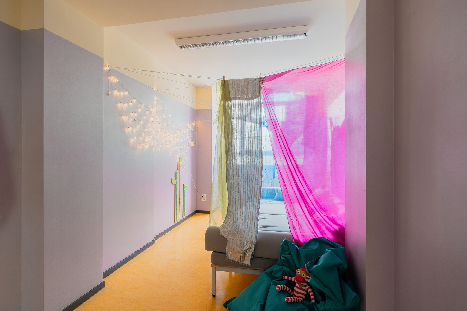 Kleiner Raum, mit leuchtender Lichterkette an der Wand. Farbige Tücher hängen vor einer Liegefläche. Durch ein Fenster fällt Licht in den Raum.