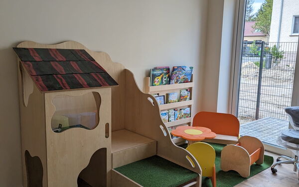Raum. Bodentiefes Fenster, Spielbereich. Mit kleiner Puppen-Sitzecke und Puppenbett. Und ei kleines Kita Kletterhaus.
