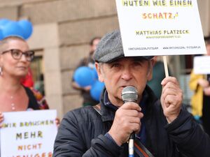 Mann mit Mikrofon in der Hand. Hält ein großes Protestschild hoch. Er trägt eine graue Schiebermütze und eine dunkelblaue Jacke. Zitat auf dem Schild: "Jede Kommune, die eine Freiwilligenagentur hat, sollte diese hüten wie einen Schatz" Matthias Platzeck