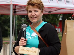 Frau mit kurzen braunen Haaren. Sie hält ein Mirkro in der rechten Hand und einen beschrifteten Holzklotz in der linken Hand vor sich. Auf dem Holzklotz steht das Wort Hilfsbereitschaft. Die Frau trägt ein türkises T-Shirt mit Aufdruck: DIE ANDERE