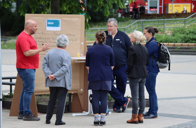 Draußen. 6 Personen stehen um einen Flipchart herum. 4 Politikerinnen unterhalten sich. 2 Männer stehen neben dem Flipshart. Einer im roten T-Shirt hält einen Stift in den Händen. Von weiterweg fotografiert.