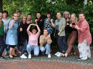 14 Frauen im Gruppenbild. Draußen vor Sträuchern. Die Frauen haben dynamische Posen eingenommen, geballte Fäuste und lächeln fröhlich und zuversichtlich.