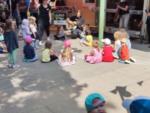 Draußen voe einem Gebäude. Kinder und Erwachsene sitzen auf dem Boden und stehen rum. Ein Mann mit Gitarre hockt bei den Kindern. Es wird musiziert.
