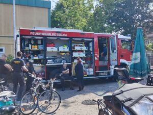 Offenes Einsatzfahrzeug der Feuerwehr Potsdam von der Seite. Einsatzkräfte stehen beim Feuerwehrauto. Davor viele geparkte Fahrräder.
