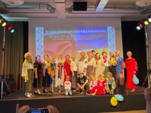 Eine Bühne mit vielen verkleideten Darsteller mit farbenfrohen Kleidern, einige tragen Blumenkränze im Haar, Kinder, Jugendlich, Erwachsene, größtenteils Frauen.