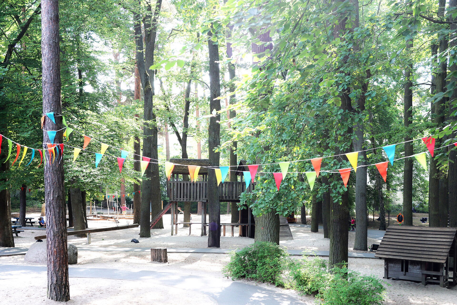 Viele Bäume durch die die Sonnenstrahlen dringen. Zwischen den Bäumen ein Spielplatz mit Wippe, Klattergerüst und vielen Bänken, alles aus Holz.