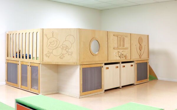 Holz gefertigte Spiel- und Kletterlandschaft für Kinder. Verschiedenen Elementen: einer erhöhten Spielfläche mit Geländer, unterschiedlichen Staufächern, einer kleinen Rutsche und an der Wand.