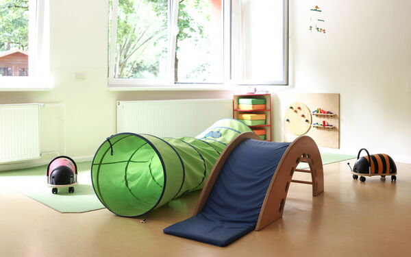 Helles Spielzimmer. Im Vordergrund befinden sich ein grüner Kriechtunnel, eine kleine blaue Rutsche aus Holz und zwei Spielzeuge, die wie Käfer aussehen.
