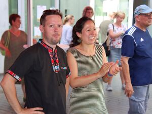 Mann und Frau stehen nebeneinander. Der Mann sieht in die kamera, die Frau klatscht und lächelt breit. Die Frau ist schlank und trägt ein ärmelloses gestreiftes Kleid, Mann mit schwarzem T-Shirt. Andere Besucher stehen rum.