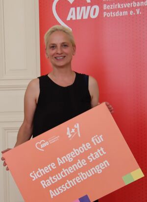 Portrait. Junge Frau mit kurzen blonden Haaren und Ohrsteckern. Sie lächelt freundlich in die Kamera. Sie hält ein großes Schild vor sich. Eine der 1+9 Forderungen. Darauf steht: Sichere Angebote für Ratsuchende statt Ausschreibungen