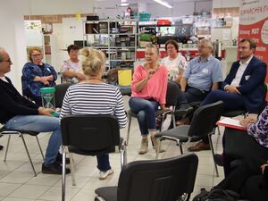 In einem Ladenraum. Zwei Stuhlkreise. Im inneren Stuhlkreis sitzen zwei Frauen und ein Mann, der Mann hält eine grüne Sanduhr auf dem Schoß. 7 Menschen sitzen im äußeren Kreis.