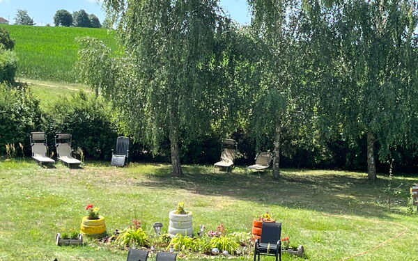 Draußen, Wiese, drei Bäue spenden Schatten an einem sonnigen Tag. Liegestühle stehen auf der Wiese. In der Mitte ein Blumenbeet.