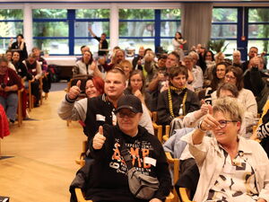 Voller Saal mit Teilnehmenden. Ein Mittelgang ist frei geräumt. Einige der Teilnehmenden lächeln und strecken den Daumen-Hoch.