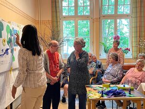 Ein Raum, große Fenster. Ältere Menschen sitzen auf Stühlen. Zwei ältere Menschen reden vor miteinander. Eine Frau mit langen dunklen Haaren gestaltet ein Plakat, diese Frau ist von hinten fotografiert.