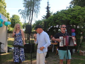 Eine Band sorgt für Stimmung. Frau in blau-weißen Sonnerkeid und 2 Männer machen Musik.