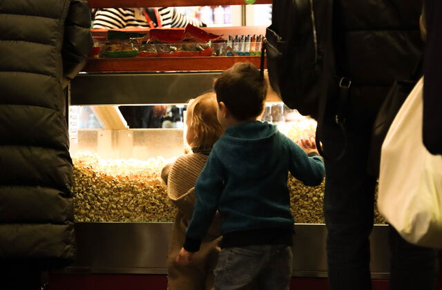 Zwei kleine Kinder stehen vor einem Tresen an dem Popcorn verkauft wird. Von hinten fotografiert. Junge in blauem Pullover/Jacke mit Kapuze. Er hat die Hand an er Scheibe das Popcorn-Verkaufs.