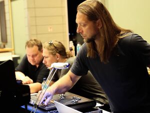 3 Männer an der Ton-Technik. Schlanker Mann mit langen glatten braunen Haaren und Bart bedient ein Mischpult. Die anderen beiden sitzen daneben.