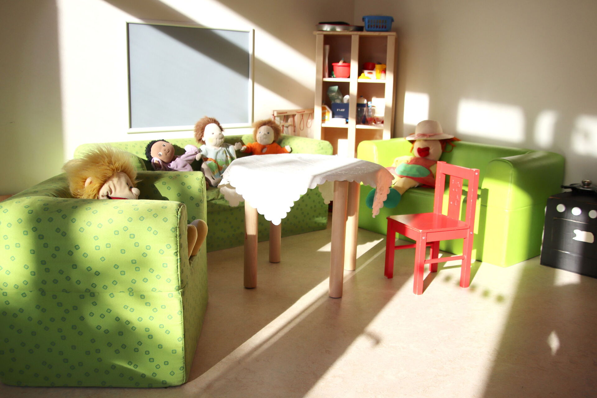 Stoffpuppen auf zwei grünen Kindersofas und einem Sessel. Ein roter Kinderstuhl steht an einem kleinen Tisch mit weißem Tischtuch in der Mitte.