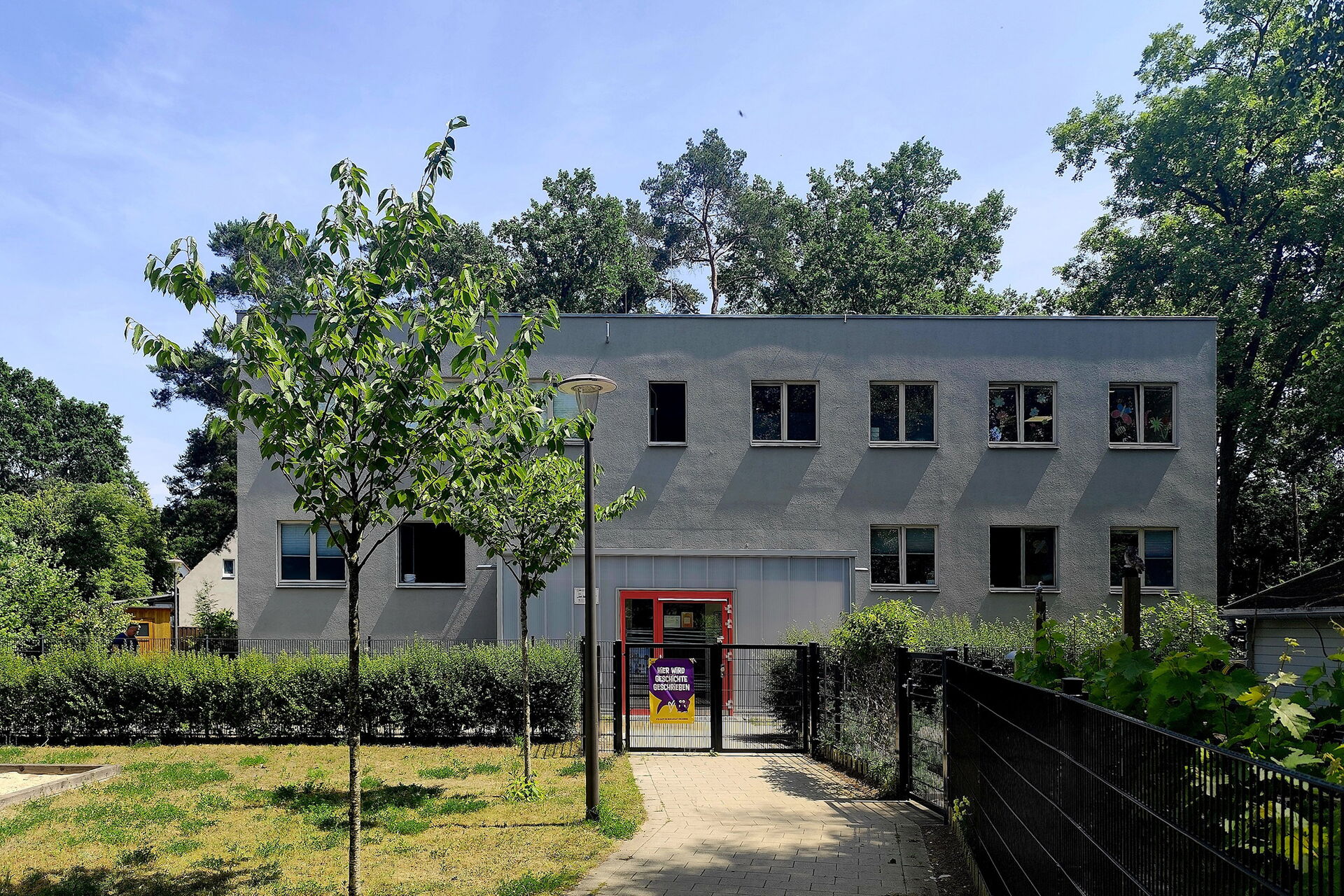 Zweistöckiges graues Gebäude. Mit einem verschlossenen Eingangstor. Vor den Gebäude: Wiese, Bäume und Hecke am Zaun.