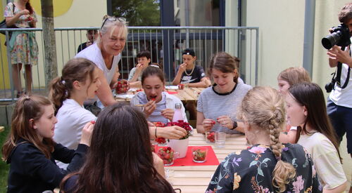 Draußen. Kinder sitzen an Holztischen.Sie essen Kirchen. Ein Fotograf fotografiert die Szene, er steht rechts neben einem Tisch. Eine frau mit blonden Haaren, Ponny, Brille auf den Kopf geschoben steht am Tisch 8 Mädchen drum herum sitzen.