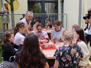 Draußen vor einem Gebäude. Kinder sitzen an Tischen und essen gut gelaunt Erdbeeren. Eine blonde Frau mit blonden Haaren und Brille auf den Kopf geschoben, steht am vorderen Tisch und lächelt.