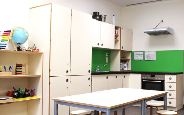 Eine Kinderküche mit großen Esstisch, Hockern, viel Stauraum durch Schränke, Herd, Abzugshaube und Spüle. Helle Möbel