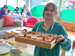 Hinten Tische wo Menschen sitzen. Im Vordergrund eine Frau mit einem Tablett mit Smoothies.