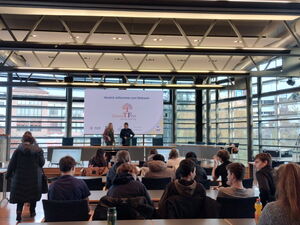 Großer verglaster Saal. Menschen sitzen an Tische. Leinwand vorne Begrüßungstext: "Herzlich willkommen zum Netzwerk Gewalt Frei."