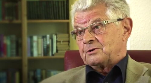 Portrait. Älterer Mann sitzt in einem Sessel. Hinter im ein Regal mit Büchern. Mann hat weiße kurze Haare, er trägt eine Brille mit silbernen Gestell. Er trägt ein dunkelblaues Hemd mit Kragen, darüber eine beigefarbene Anzugjacke.