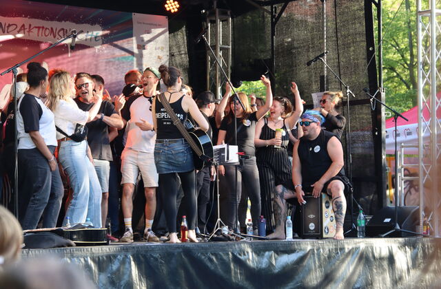 Große Gruppe von Menschen auf der Bühne beim Singen. Eine Frau mit Gitarre und ein Mann trommelt den Rhythmus,