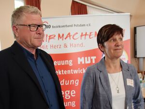 Ein Mann und eine Frau stehen vor einem großen AWO Aufsteller. Mann blonde kurze Haare, Brille mit schwarzen Rahmen, dunkles Hemd, schwarzes Sakko, die Frau hat braune kurze Haare weißes T-Shirt unter langärmliger Bluse.