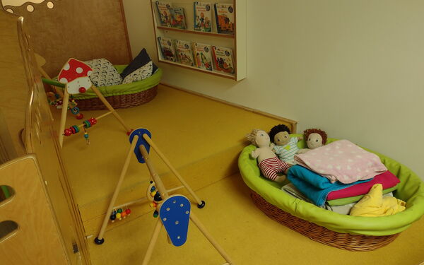 Zwei Kleinkindkuschelnester mit Decken und Puppen als Inhalt und zwei Spielbögen auf gelbem Teppich