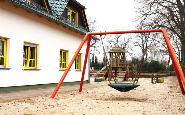 Rechts weißes Haus mit gelben Fensterrahmen und grünem Dach. Spielsplatz mit Vogelnestschaukel und dahinter ein Klettergerüst mit Rutsche.