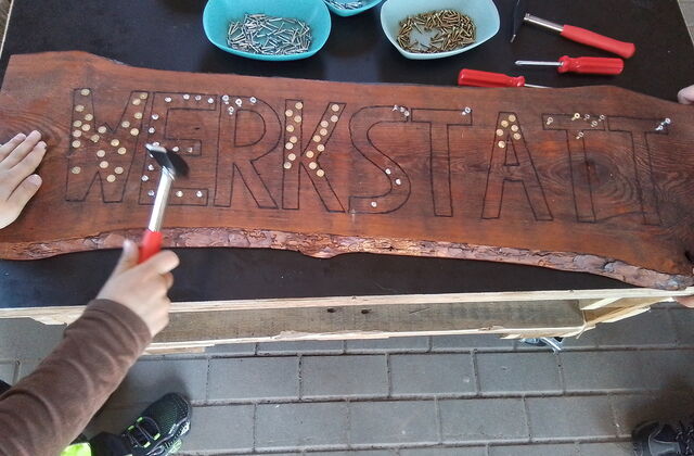 Großes Holzschild auf einer Werkbank. Auf dem Schild steht in Großbuchstaben das Wort Werkstatt.      Kinder füllen die Buchstaben mit Reiszwecken, Kind mit Hammer links im Bild.