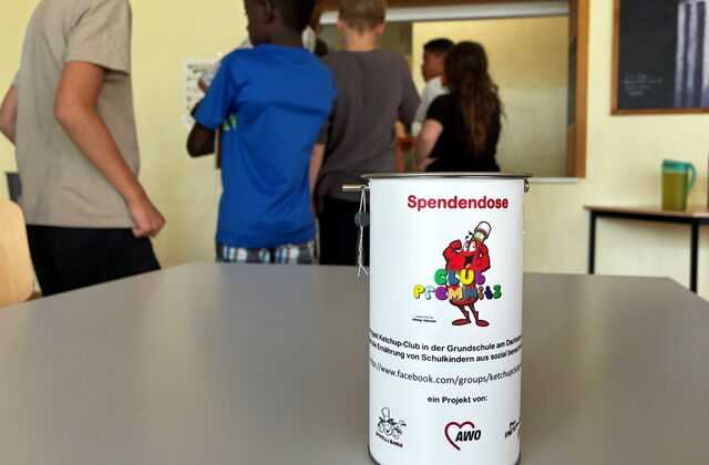 Im Vordergrund eine weiße Spendendose mit Logo vom Ketchup Club. Im Hintergrund Kinder.