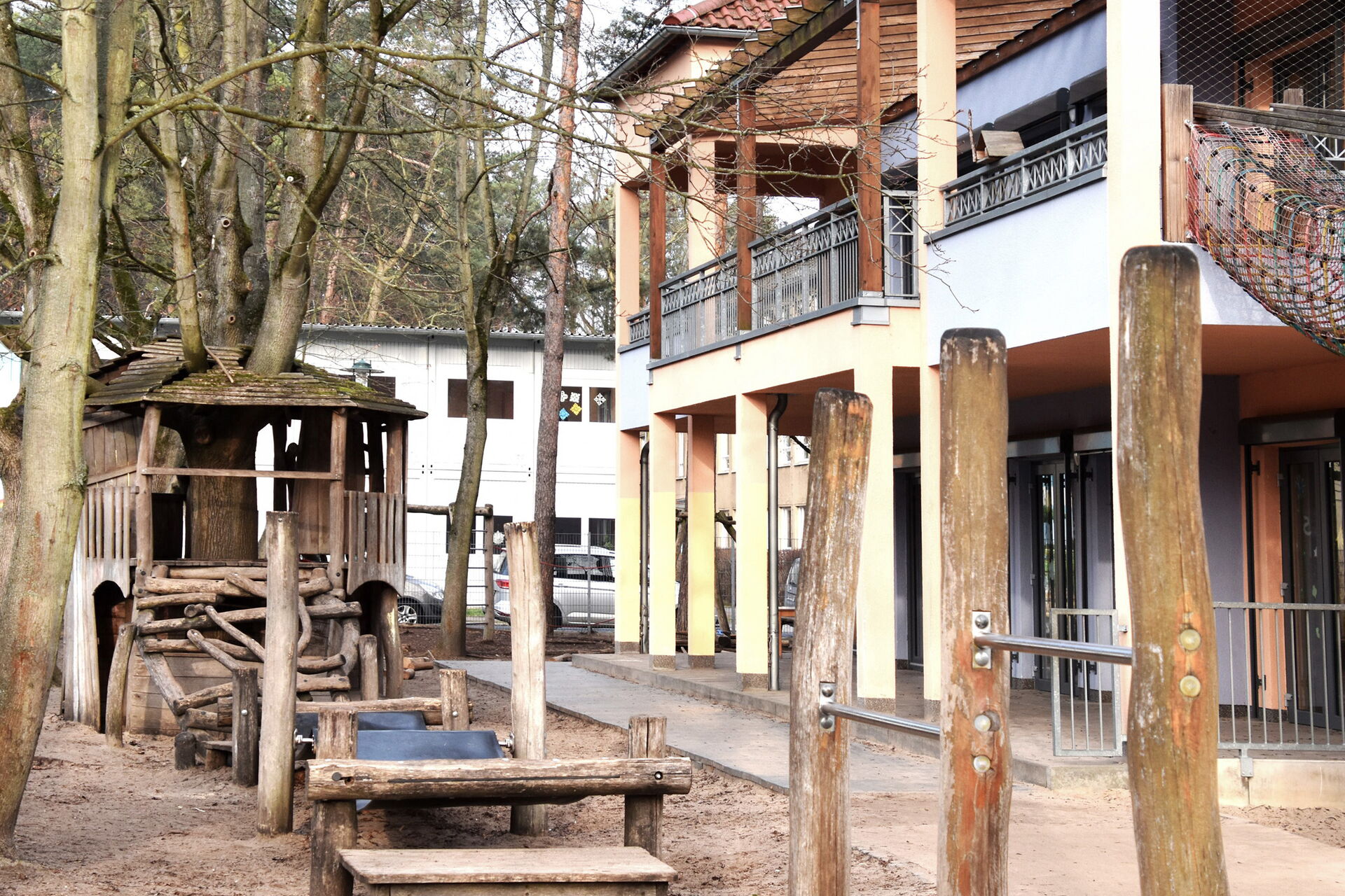 Draußen, Bänke und Tische aus Baumstämmen, Holz-Klettergerüst. Rechts daneben ein Weg und das Kita Gebäude, im zweiten Stock mit Balkon.
