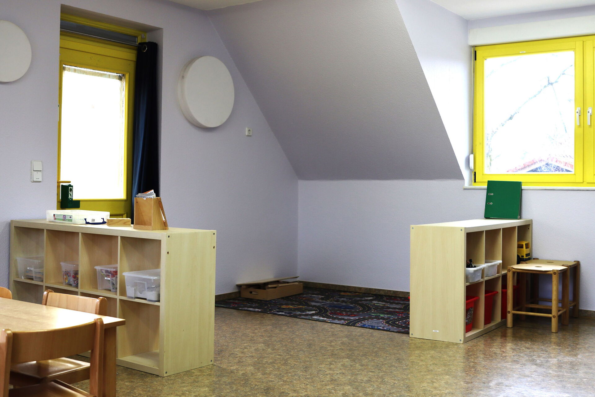 Spielzimmer. In der Ecke liegt ein Straßenteppich. Zwei kleine Regale stehen bei den Fenstern. Ein kleiner Tisch mit kleinen Stühlen.