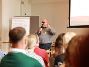 Teilnehmende sitzend von hinten. Vorne ein korpulenter Mann in grauen Hemd mit Mikrofon in der Hand.