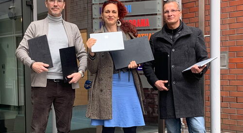 2 Männer in der Mitte eine Frau, alle drei halten Laptops und Notebooks in den den Händen. Sie stehen vor dem Gebäude der UFA.