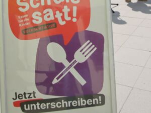 Draußen, Sonne scheint. Aufsteller Schild Text: Schule satt! Essen für alle Kinder. Jetzt unterschreiben.       Dahinter drei Personen. 2 Frauen, ein Mann in der Mitte am Mikro.