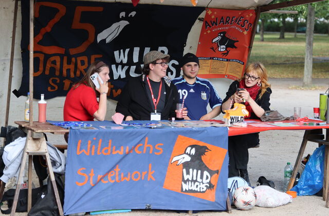 Stand. Mit Banner 25 Jahre Wildwuchs. 4 Menschen sitzen an dem Stand.