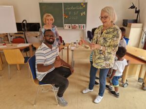 Ein Klassenzimmer. Zwei weiße Frauen stehen im Raum. Auf einem Stuhl sitzt ein scharzer Mann. Sie lächeln in die Kamera und mach die Daumen hoch Geste.