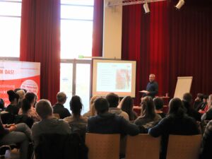 Großer Raum, viele Menschen sitzen auf Stühlen von hinten. Eine Leinwand wird mit Beamer bespielt. Vorne steht ein Redner.