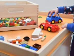 Spieletisch mit Lego und keinem blauen Auto. Eine Kinderhand umfasst die Kante des Tisches mit der anderen Hand schiebt es das Auto.