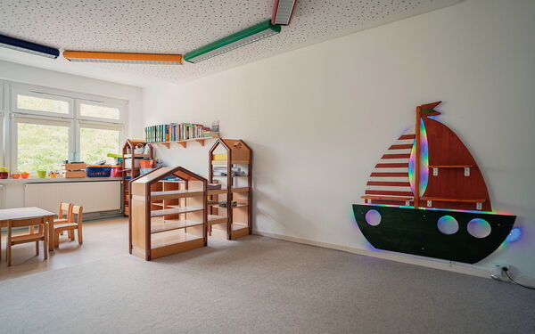 Großer heller Raum, hellgrauer Teppich im ganzen Raum. An der weißen Wand ein beleuchtetes Segelschiff aus Holz. Kleine Regale, eine Spielecke mit kleinem Tisch und Stühlchen.
