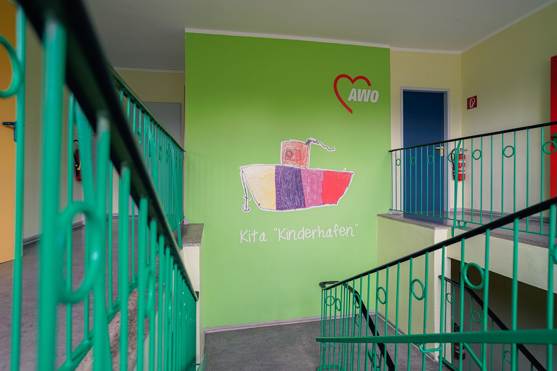 Treppenhaus in Grün. Auf einer Wand das AWO Logo und ein buntes Schiff mit Anker. Unter dem Schiff steht in weißer Schrift Kita "Kinderhafen".