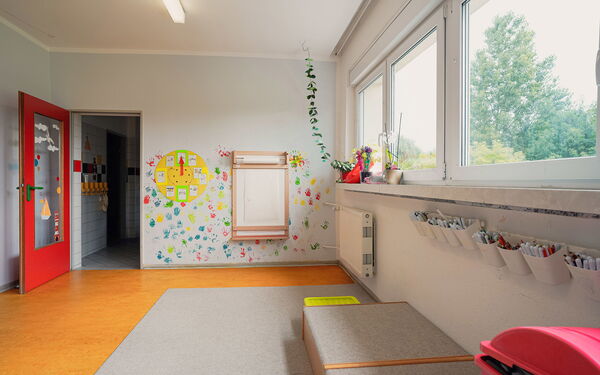Raum, rote Tür steht offen. Im Raum ist ein hellgrauer Teppich. An der Wand neben der roten Tür ist eine gebastelte Uhr, und an der Wand sind farbige Handabdrücke der Kinder in blau, gelb, rot und grün.
